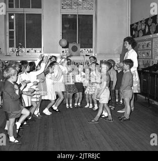 1960s, historische Grundschulklasse, eine Lehrerin, die eine Gruppe von kleinen Kindern überwacht, die mit einer kleinen Lichtkugel im Klassenzimmer spielen. Stockfoto