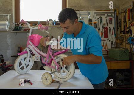 Bild eines jungen Handwerkerdades, der in seiner Werkstatt das Fahrrad seiner Tochter repariert. Do-it-yourself-Arbeit Stockfoto