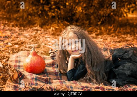 Halloween. Ein süßes Mädchen mit langen Haaren liegt auf einer orangefarbenen Decke im Park neben einem lustigen Kürbis. Stockfoto