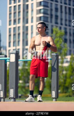 Schöner junger muskulöser Mann in roten Shorts auf dem Sportplatz im öffentlichen Park am Sommermorgen. Sport, Fitness, gesundes Lifestyle-Konzept Stockfoto
