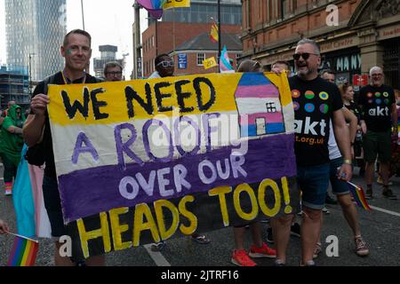 Manchester Pride Parade. AKT Obdachlose Wohltätigkeitsorganisation marschiert mit Bannertext Wir brauchen auch ein Dach über unseren Köpfen. Themenmarsch für den Frieden. Stockfoto