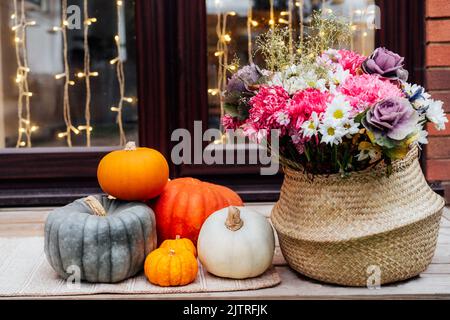 Haus für Herbstferien dekoriert. Tür beleuchtet mit Girlande und Veranda mit verschiedenen Kürbissen und Blumen im Korbkorb für Thanksgiving o Stockfoto