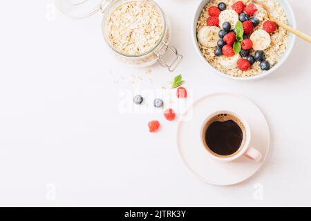 Kaffee und hausgemachter Haferbrei mit Himbeeren, Heidelbeeren, Banane und Chia-Samen auf weißem Tisch. Gesundes Frühstück. Draufsicht, flach liegend, Kopierbereich. Stockfoto