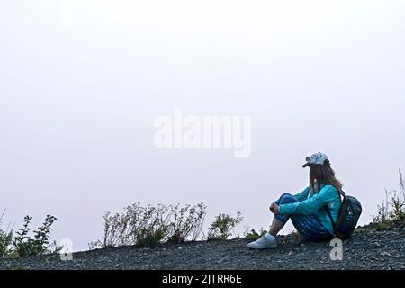 Junge Frau in Mütze und blaue sportliche Kleidung mit Rucksack sitzt am Rande des Berges gegen weiße Wolken und Nebel Blick Tourismus Wandern und ein Stockfoto