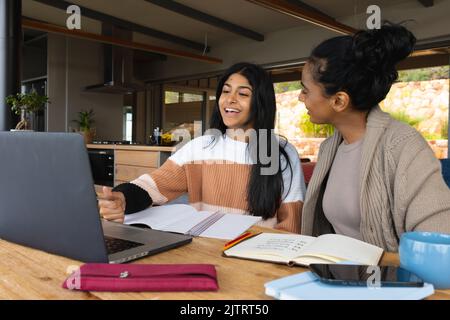 Birazialmutter, die zu Hause eine glückliche Teenager-Tochter ansieht, die über einem Laptop auf dem Tisch studiert Stockfoto