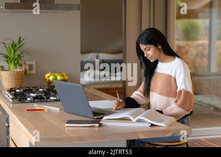 Biracial fokussiertes Teenager-Mädchen, das in einem Buch schreibt, während es mit einem Laptop auf der Kücheninsel zu Hause sitzt Stockfoto