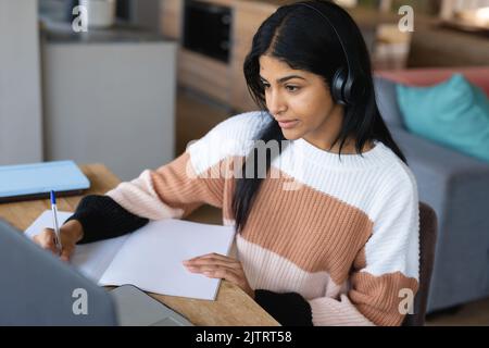 Biracial Teenager Mädchen trägt Kopfhörer und schriftlich in Buch, während der Teilnahme an Online-Vortrag Stockfoto