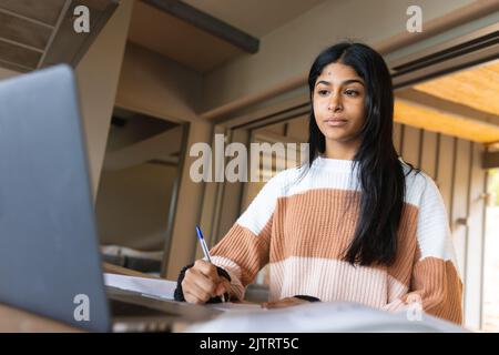 Biracial serious Teenager-Mädchen mit langen Haaren schriftlich in Buch und Blick auf Laptop auf dem Tisch zu Hause Stockfoto