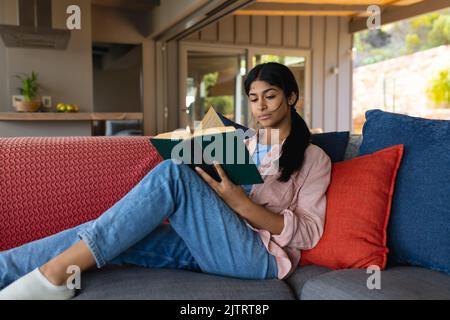 Biracial fokussierte Teenager-Mädchen mit langen Haaren Buch lesen, während auf der Couch im Wohnzimmer entspannen Stockfoto