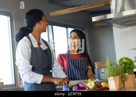 Lächelnde Birazialmutter und Tochter, die einander angucken, während sie Gemüse in der Küche hacken Stockfoto