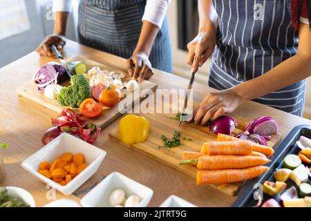 Mittelteil der birazialen Mutter und Tochter trägt Schürzen und hackt frisches Gemüse auf dem Tisch Stockfoto