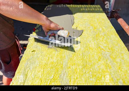 Blick auf die Hand der Arbeiter mit Kelle, Aufbringen von Klebstoff auf gelbe Mineralwolle zur Isolierung des Gebäudes im Bau. Stockfoto