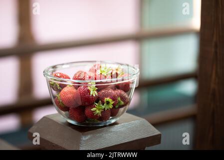 Frische, leckere Erdbeeren in einer transparenten Glasschale auf einer braunen Holzoberfläche. Unscharfer Hintergrund. Stockfoto