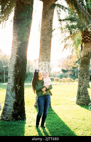 Mama sieht das Baby in ihren Armen an, während sie unter einer großen Palme steht Stockfoto