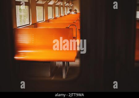 Ein Foto des Innenraums eines leeren Retro-Zuges mit orangefarbenen Sitzen an einem hellen Tag, aufgenommen durch das Glas der Tür. Pendlerzüge. Stockfoto