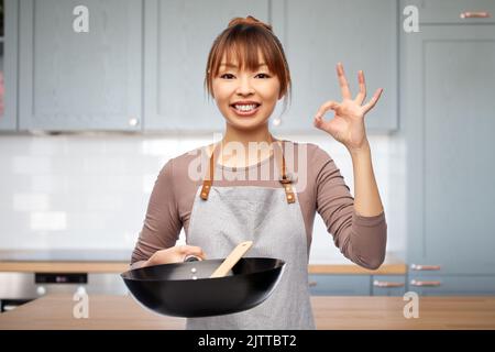 Frau auf der Schürze mit Bratpfanne zeigt in der Küche ok Stockfoto