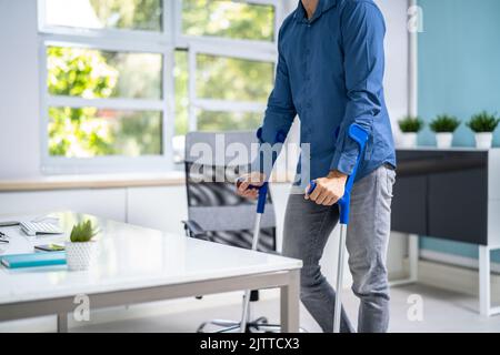 Arbeiter Mit Krücken Am Arbeitsplatz Oder Im Büro. Rehabilitationsleistungen Stockfoto