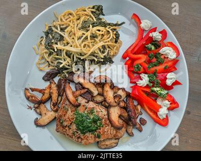 Draufsicht auf einen hellgrauen Essteller mit gebratenem Thunfisch, gerösteten Shiitake-Pilzen, Spaghetti mit Kapern und Spinatblättern und rohem roten Paprika Stockfoto