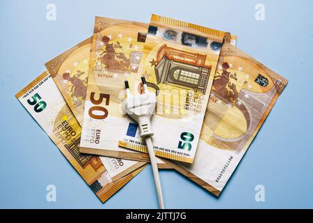 Der elektrische Stecker liegt auf den Euro-Banknoten und zeigt die drastische Inflation bei den Stromkosten. Energiekrisenkonzept in Europa. Draufsicht. Blauer Hintergrund Stockfoto