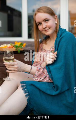Porträt einer jungen, prallen Frau, die in der Nähe von geschnittenem Obst, Gemüse und Gemüse auf einem Holztablett sitzt und ein Glas Weißwein hält. Stockfoto