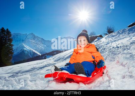 Niedlicher kleiner Junge geht im roten Schlitten in den Bergen bergab Stockfoto