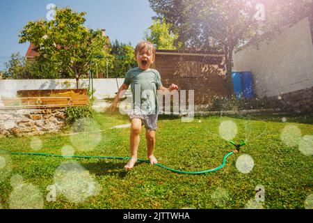 Lachend hübscher Junge läuft im Garten um den Wasserregner herum Stockfoto