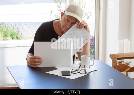 Frustrierter Mann mit Mütze, der Papierdokumente las, während er am Tisch saß. Papiere, Gläser, Becher und Handy auf dem Tisch. Stockfoto