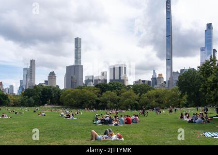 New York City, USA - 21. August 2022: Menschen entspannen sich auf dem Gras auf der Sheep Meadow des Central Park mit Gebäuden im Hintergrund in New York City. Stockfoto