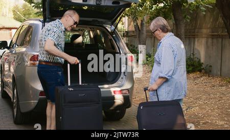 Ältere Leute, die Gepäck in den Kofferraum legen, während sie sich gemeinsam auf die Reise in den Ruhestand vorbereiten. Älteres Paar mit schwerem Reisegepäck geht auf Feldreise, um den Hochzeitstag zu feiern Stockfoto