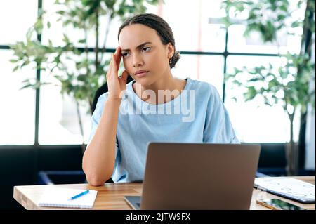 Erschöpft müde gestresst traurig gemischte Rasse junge Frau, Büroangestellte, sitzt am Schreibtisch im Büro, überarbeitet, müde von der Arbeit in einem Laptop an einem Projekt, Kopfschmerzen, braucht eine Pause, Ruhe Stockfoto