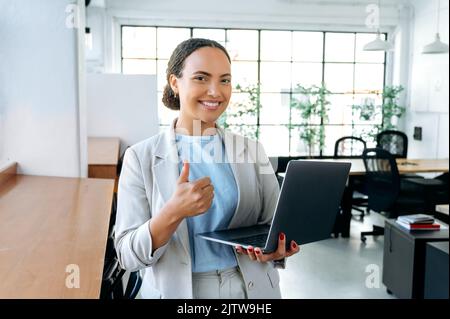 Selbstbewusste, hübsche, freundliche junge Frau mit hispanischer oder brasilianischer Nationalität, steht in einem modernen Kreativbüro, hält einen Laptop in der Hand, blickt auf die Kamera, lächelt, zeigt eine Geste des Daumen nach oben Stockfoto
