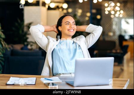 Ruhige, entspannte, gemischte Mitarbeiterin, die während der Arbeitszeit ausruht, im modernen Büro sitzt, die Hände hinter den Kopf stellt, sich mit dem Projekt zufrieden fühlt, die Arbeit befördert, in die Ferne blickt und freundlich lächelt Stockfoto