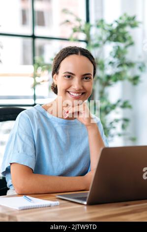 Vertikales Foto einer schönen, eleganten brasilianischen oder hispanischen jungen Frau, Maklerin, Kreativmanagerin, ceo-Firma, sitzt auf einem Schreibtisch mit Laptop in einem modernen Kreativbüro und schaut lächelnd auf die Kamera Stockfoto