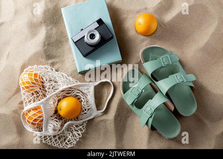Pantoffeln, Beutel mit Orangen, Kamera und Buch am Strand Stockfoto