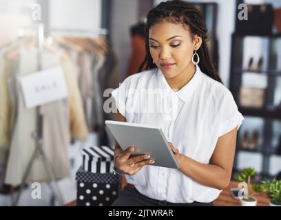 Online die neuesten Fashons im Auge behalten. Eine junge Geschäftsbesitzerin benutzt ihr Tablet, während sie in ihrem Geschäft steht. Stockfoto