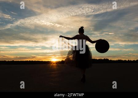 Frau, die in den Strahlen der untergehenden Sonne steht, ihre Arme an den Seiten ausgestreckt und den Rand des Tutu-Rockes und des Hutes gegen den magischen Himmel mit Wolken hält Stockfoto