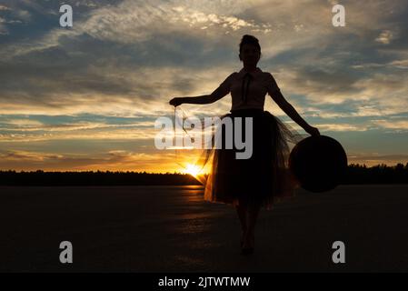 Frau hält Rand des Tutu Rock gegen Wald Silhouette und Sonnenuntergang wolkigen Himmel durch Sonnenstrahlen der untergehenden Sonne mit Hut in der Hand hinterleuchtet, niedrigen Winkel Ansicht Stockfoto