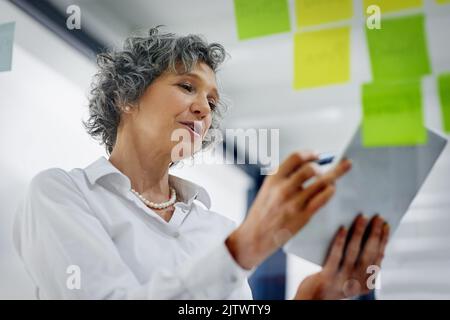 Eine reife Geschäftsfrau, die in einem modernen Büro Notizen an eine Glaswand schreibt, verwendet Technologie zur Verwaltung ihres Geschäfts. Stockfoto
