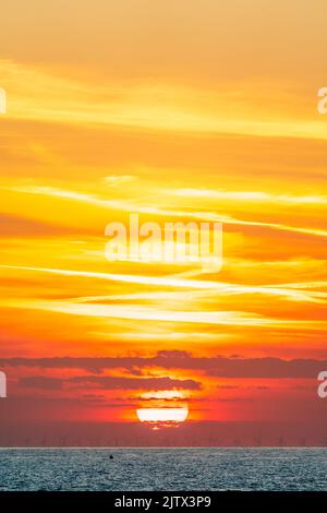 Die Sonne, die hinter einer Wolkenschicht am Horizont über der Nordsee vor der Küste von Kent in einen orange-gelben Himmel aufgeht. Verschiedene dünne Wolkenschichten über der Sonne, die vom Sonnenlicht hell erleuchtet ist. Stockfoto