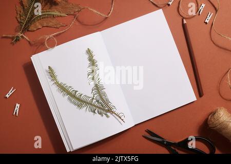 Getrocknetes gepresstes Blatt auf Papier aus Notizbuch mit Bleistift, Schere und Garn auf braunem Hintergrund. Hobby, handgemachte, florale Kunst und Boho Style Konzept Stockfoto