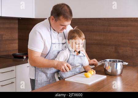 Konzept Nahrung und Ernährung. Aufnahme von zwei fröhlichen Papa und Sohn, die in der Küche posieren und Kartoffeln schälen, die zusammen vegetarisches Essen zum Abendessen kochen, mit identischen Schürzen Stockfoto