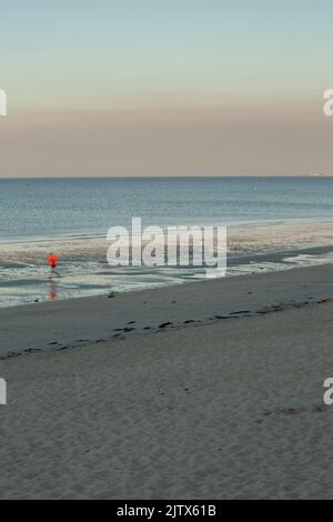 Ein Läufer in einem roten T-Shirt läuft an einem ruhigen Morgen am nassen Strand. Das Meer ist ruhig und die Farben sind pastellfarben. Stockfoto