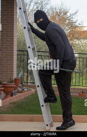 Dieb in schwarz gekleidet, mit einem Brecheisen, der eine Leiter klettert und versucht, sich in ein Haus zu schleichen Stockfoto