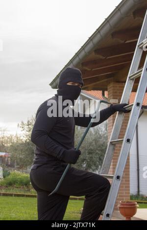 Dieb in schwarz gekleidet mit Brecheisen und Balaclava, der eine Leiter klettert, während er versucht, sich in ein Haus zu schleichen Stockfoto