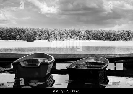 Zwei Boote in der Nähe eines hölzernen Pier mit einem Wald im Hintergrund in Graustufen Stockfoto