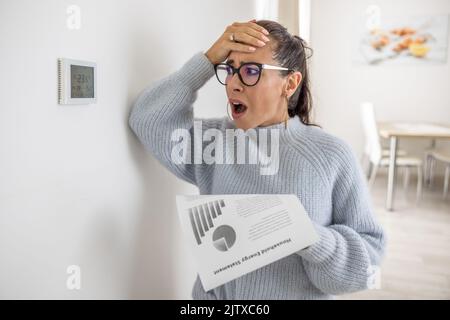 Die Frau kann ihren eigenen Augen die Summe auf ihrer Energierechnung nicht glauben und sieht schockiert über den Thermostat an der Wand neben ihr aus. Stockfoto