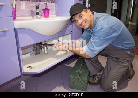 Bild eines lächelnden Klempners, der Rohre an einem Waschbecken repariert. Sanitär- und Heimwerkerarbeiten. Stockfoto