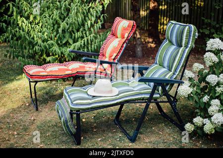 Zwei weiche Sonnenliegen mit Strohhut im Innenhof. Zwei leere Sonnenliegen in einem sonnigen Garten zur Entspannung. Stockfoto