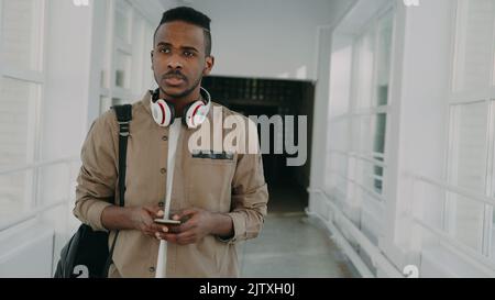 Hübscher afroamerikanischer Student mit großen weißen Kopfhörern, der in einem langen, hellen Korridor der Hochschule mit einem Smartphone unterwegs ist, das jemandem eine SMS schreibt Stockfoto