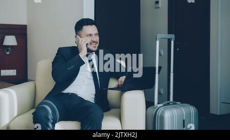 Selbstbewusster, fröhlicher Geschäftsmann im Anzug, der den Laptop überprüft und am Mobiltelefon spricht, während er im Sessel im Hotelzimmer sitzt. Business-, Reise- und Mitarbeiterkonzept Stockfoto
